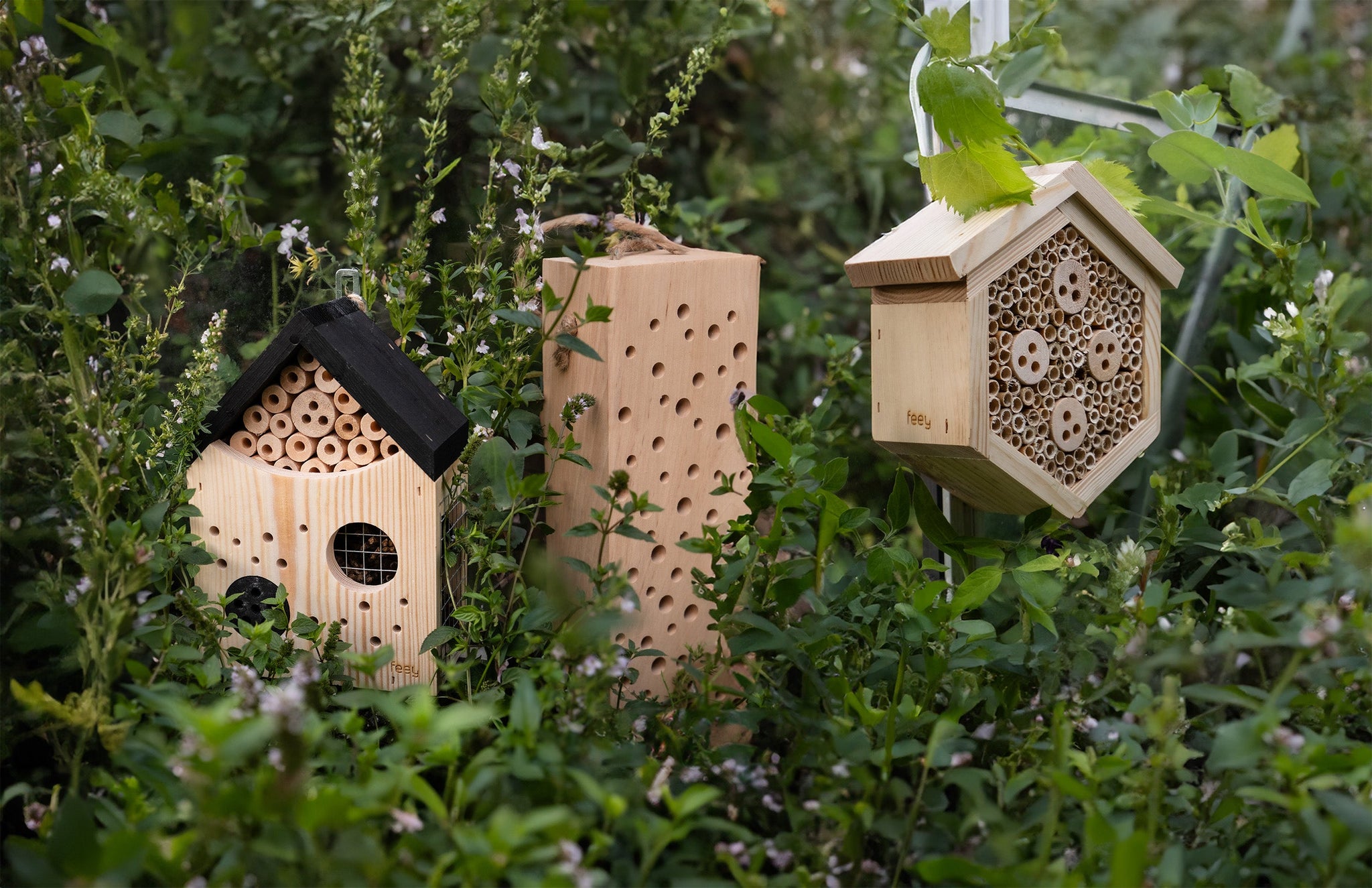 Drei hölzerne Bienenhäuser in unterschiedlichen Formen stehen inmitten üppiger grüner Blätter. Die Häuser verfügen über mehrere kleine Löcher, in denen Bienen Platz finden, und verleihen der Gartenumgebung eine natürliche Ästhetik.