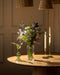 Der Strauß "Purpurpoesie" aus violetten gebundenen Blumen in einer Glasvase schmückt einen runden Holztisch, begleitet von weißen Blüten in einer grünen Vase - ein Arrangement, das Eleganz und Poesie ausstrahlt, ideal als Blumengeschenk bei warmem Licht.