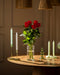Ein Klassiker: Rote Rosen (Inventar) in klarer Glasvase auf rundem Holztisch, umgeben von grünen und weißen Kerzen in goldenen und gläsernen Haltern unter sanftem Licht - eine Liebeserklärung.