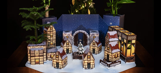 Eine festliche Szene mit von innen beleuchteten Miniatur-Papierhäusern, die an ein verschneites Dorf erinnern. Kleine Bäume und ein Sternenhimmel im Hintergrund verstärken die gemütliche, winterliche Atmosphäre.