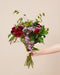 Eine Hand hält den Amore-Strauss mit dunkelroten Alstroemerien, rosa Statice-Blüten und grünen Gräsern vor einem warmen beigen Hintergrund.