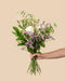 Eine Hand hält den Blumenstrauß "Wolkige Grüße" mit weißen Rosen, pinken Knospen, gelben Akzenten, violetten Blüten und grünen Blättern vor beigem Hintergrund - eine wunderschöne Geschenkidee.