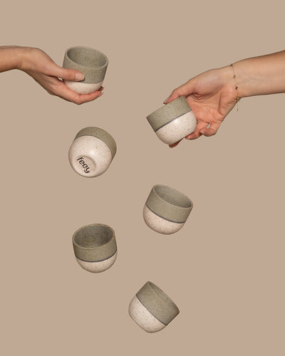 Zwei Hände halten Keramiktassen vor einem beigen Hintergrund. Vier weitere Teile aus dem Keramik Baby-Topfset 'Variado' (6 Stk.) scheinen zwischen ihnen zu schweben. Die Tassen haben ein gesprenkeltes Design mit hellerer Unterseite und dunklerer Oberseite. Ein Babytopf trägt auf der Unterseite ein Logo mit dem Text „freey“.