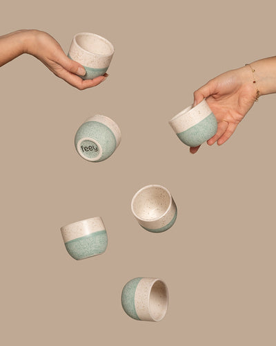 Zwei Hände greifen nach fallenden Keramiktassen vor einem beigen Hintergrund und präsentieren das Keramik Baby-Topfset „Variado“ (6 Stk.). Diese Töpfe haben ein gesprenkeltes Design mit grünem Boden und cremefarbenem Deckel, und einer zeigt das Wort „Feed“ auf der Unterseite. Die Szene strahlt Eleganz und Bewegung aus.