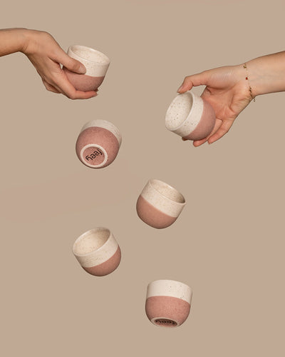 Zwei Hände halten rosa-weiß gesprenkelte Tassen aus dem Keramik Baby-Topfset 'Variado' (6 Stk.), während vier weitere Tassen aus demselben Set in der Luft zu schweben scheinen. Der einfarbig beige Hintergrund verleiht der Szene eine ruhige Note.