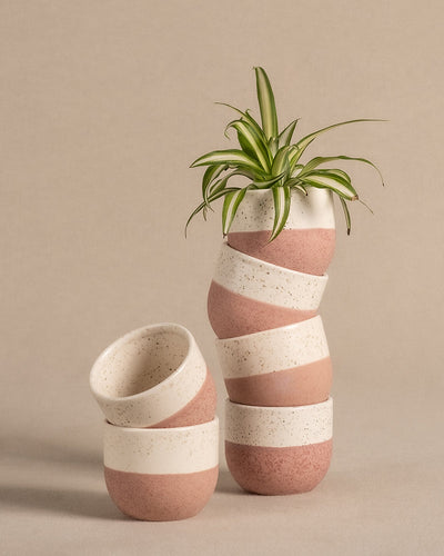 Das Keramik Baby-Topfset 'Variado' (6 Stk.) besteht aus einem Stapel von fünf Keramiktöpfen mit abwechselnd rosa und weißen Streifen, gekrönt von einer leuchtend grünen Pflanze. Der neutrale beige Hintergrund ergänzt die verspielten Farben der Töpfe perfekt.