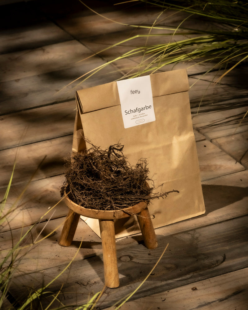 Ein kleiner orange-brauner Beutel mit der Aufschrift „Schafgarbe (orange-braun)“ steht hinter einem Holzhocker mit getrockneten Kräutern, der auf einem Holzboden mit verstreuten Pflanzenschatten steht.