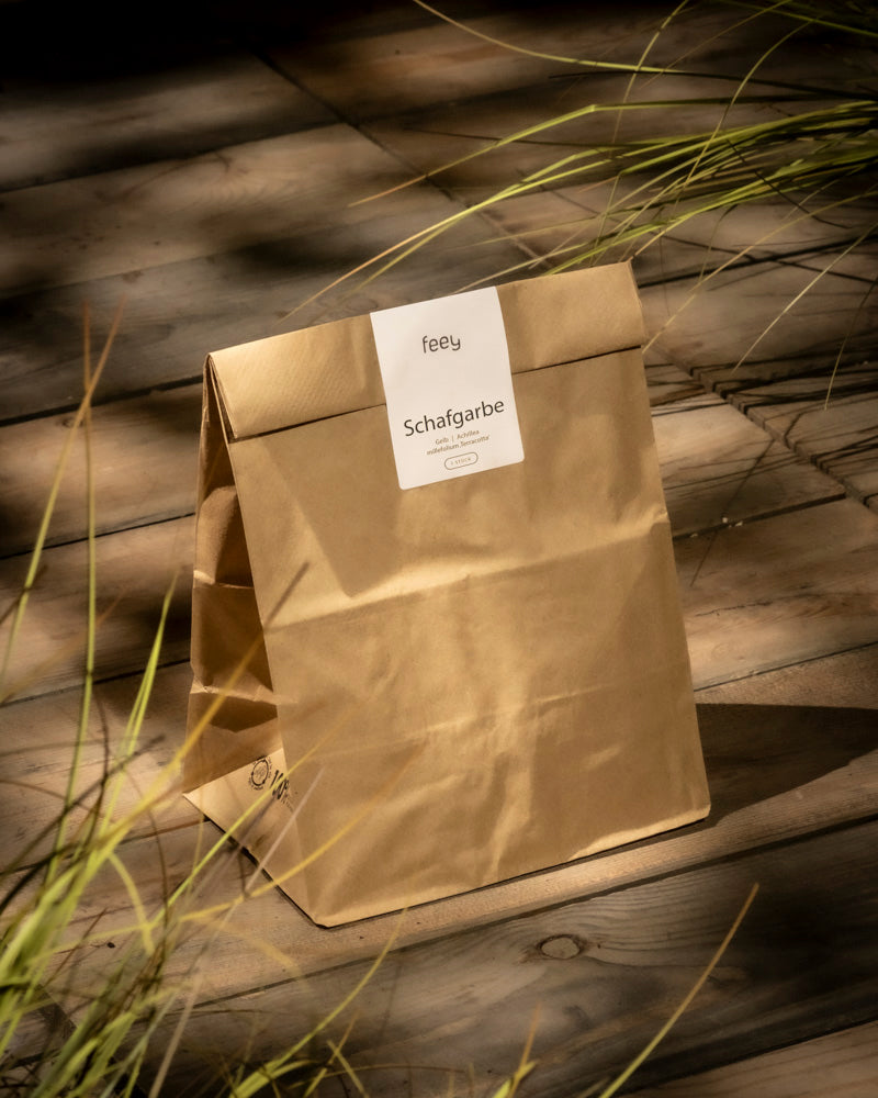 Eine braune Papiertüte mit der Aufschrift „Schafgarbe (orange-braun)“ liegt auf einer sonnenbeschienenen Holzoberfläche, teilweise eingerahmt von hohem grünen Gras und präsentiert das Kraut Achillea millefolium.