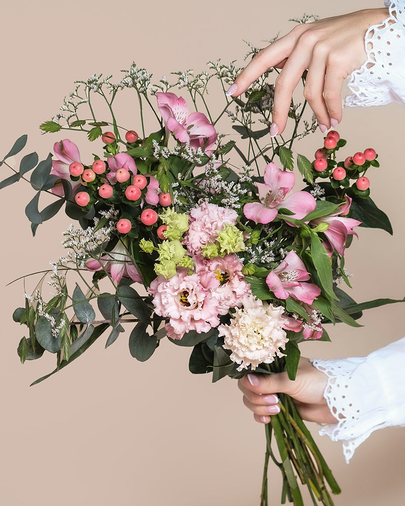 Eine Hand hält den Ballerina-Strauß - rosa und weiße Blumen, grüne Blätter und rote Beeren - vor einem beigen Hintergrund, während eine andere Hand mit Spitzenmanschetten die Blüten sanft von oben arrangiert.