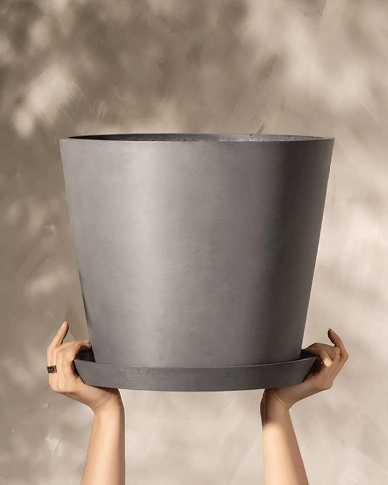 Zwei Hände, die aus dem unteren Bildrand hervortreten, halten einen großen, leeren grauen Kunststoff-Pflanzkübel (Amsterdam | 40 cm ⌀) von EcoPots vor einem neutralen Hintergrund mit subtilen Schatten. Die Hände liegen leicht geneigt am Boden des Topfes, die Finger sind zur Unterstützung gespreizt.