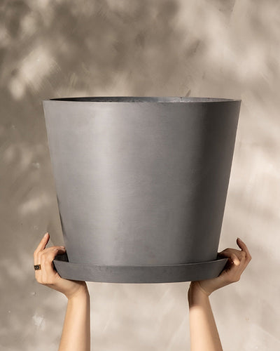 Ein Paar Hände hält einen großen, leeren, grauen Kunststoff-Pflanzkübel (Amsterdam | 40 cm ⌀) mit passendem Sockel vor einem neutralen, strukturierten Hintergrund. Geflecktes Licht fällt auf die Szene und erzeugt weiche Schatten, die das integrierte Drainage-System hervorheben.
