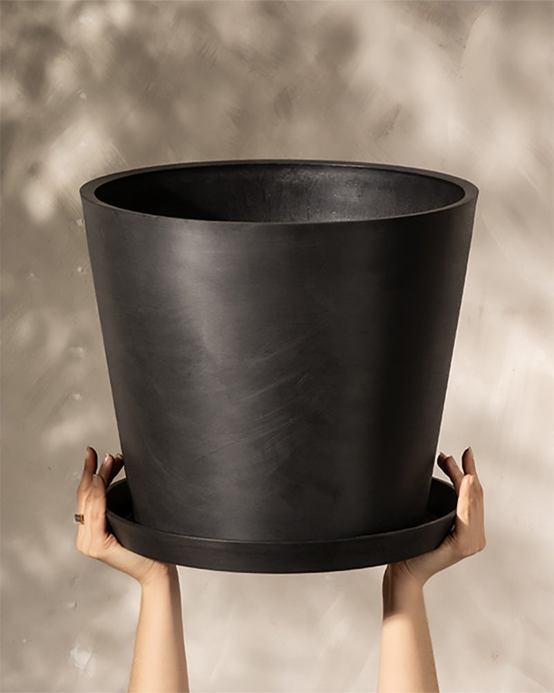 Zwei Hände halten einen großen, mattschwarzen Kunststoff-Pflanzkübel (Amsterdam | 40 cm ⌀) mit passendem Untersetzer hoch. Der Hintergrund ist eine unscharfe hellbeige Wand mit weichem, gesprenkeltem Licht. Dieser Kunststoff-Pflanzkübel (Amsterdam | 40 cm ⌀) verfügt außerdem über ein effizientes Drainagesystem, damit Ihre Pflanzen gesund bleiben.
