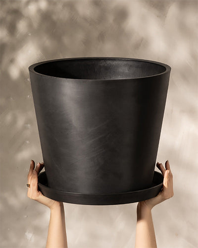 Zwei Hände halten einen großen, mattschwarzen Kunststoff-Pflanzkübel (Amsterdam | 40 cm ⌀) mit passendem Untersetzer hoch. Der Hintergrund ist eine unscharfe hellbeige Wand mit weichem, gesprenkeltem Licht. Dieser Kunststoff-Pflanzkübel (Amsterdam | 40 cm ⌀) verfügt außerdem über ein effizientes Drainagesystem, damit Ihre Pflanzen gesund bleiben.