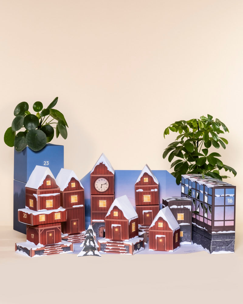 Der Pflanzen-Adventskalender hat die Form eines verschneiten Dorfes mit beleuchteten Fenstern und nummerierten Türen, flankiert von zwei grünen Blumentöpfen mit echten Pflanzen, alles vor einem beigen Hintergrund.