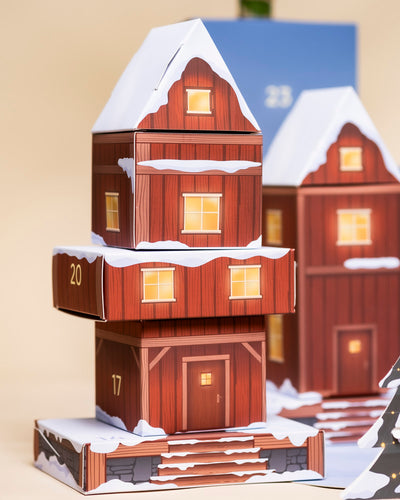 Eine Nahaufnahme des festlichen Pflanzen-Adventskalenders (Dschungel-Version), der die Form eines verschneiten Holzhauses hat. Leuchtend gelbe Lichter leuchten durch die Fenster, die Türen 17 und 20 sind sichtbar und deuten auf Zimmerpflanzen-Überraschungen im Inneren hin.