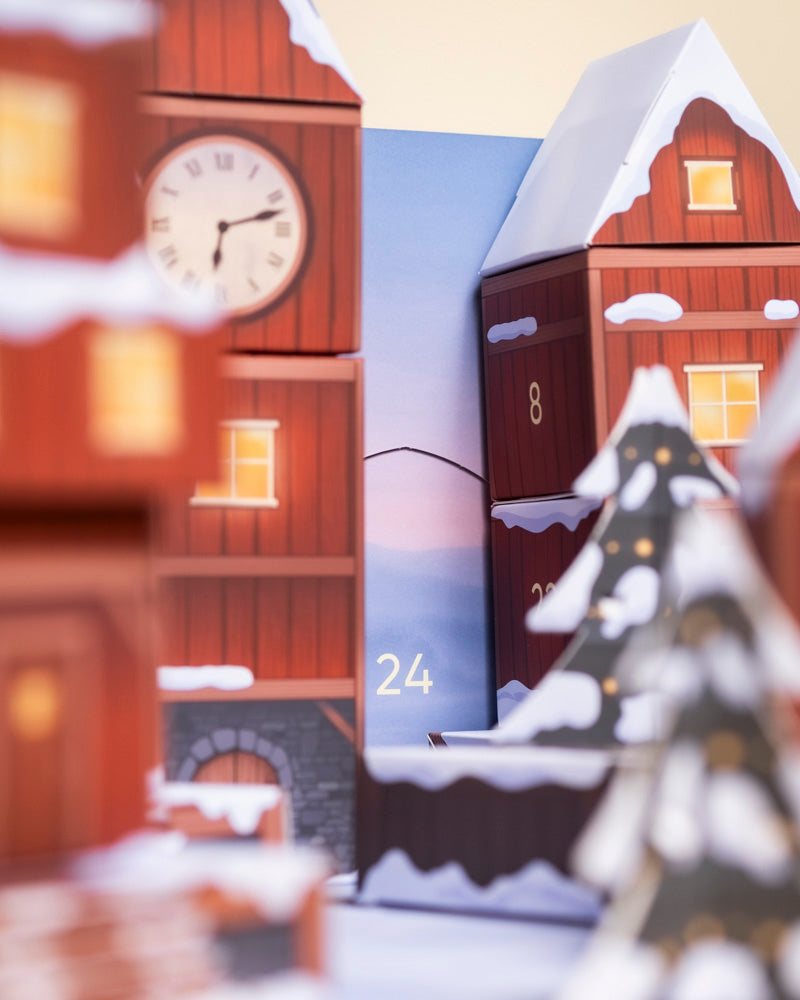 Eine Nahaufnahme einer festlichen Szene mit roten Holzhäusern, verschneiten Dächern, einer Uhr und einem geschmückten Baum. Die Zahl "24" weist auf den Pflanzen-Adventskalender (Dschungel-Version) vor einem bezaubernden pastellfarbenen Himmelshintergrund hin.