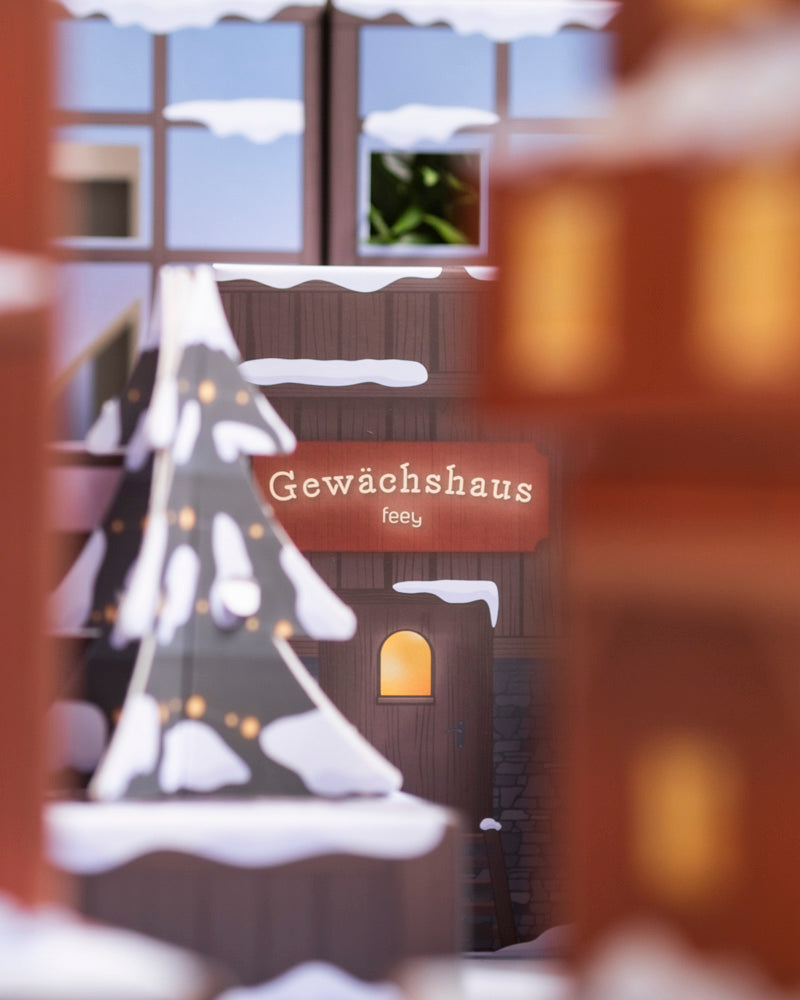 Durch ein Fenster mit einem "Gewächshaus feey"-Schild sieht man ein gemütliches, verschneites Gewächshaus. Drinnen leuchtet ein Weihnachtsbaum inmitten üppiger Zimmerpflanzen - perfekt für Fans des Pflanzen-Adventskalenders (Dschungel-Version).