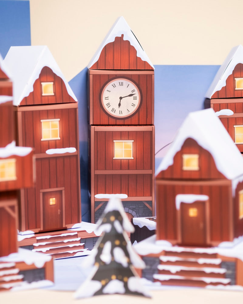 Der Pflanzen-Adventskalender zeigt eine festliche Winterszene: schneebedeckte rote Häuser, leuchtende Fenster, ein 2:15-Uhr-Turm und echte Topfpflanzen neben einem geschmückten Weihnachtsbaum sorgen für Weihnachtsstimmung.