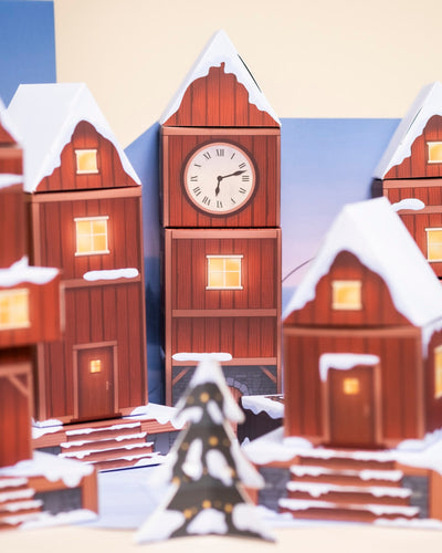 Der Pflanzen-Adventskalender zeigt eine festliche Winterszene: schneebedeckte rote Häuser, leuchtende Fenster, ein 2:15-Uhr-Turm und echte Topfpflanzen neben einem geschmückten Weihnachtsbaum sorgen für Weihnachtsstimmung.