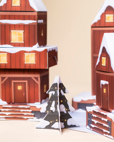 Eine Nahaufnahme des Pflanzen Adventskalenders zeigt ein verschneites Pappdorf mit roten Häusern, einem festlichen Weihnachtsbaum, leuchtenden gelben Lichtern und kleinen Pflanzentöpfen, die eine bezaubernde Weihnachtsszene schaffen.