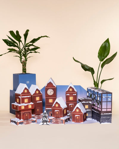 Der Pflanzen-Adventskalender zeigt kleine schneebedeckte Modellhäuser mit nummerierten Türen, die zwischen echten Topfpflanzen eingebettet sind und eine bezaubernde Winterdorfszene vor einem beigen Hintergrund bilden.