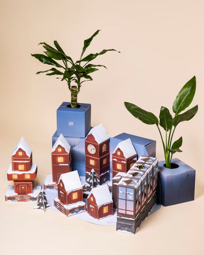Eine festliche Winterdorfszene mit Papierhäusern und schneebedeckten Bäumen umgibt den blauen Pflanzen-Adventskalender mit echten Pflanzen in Töpfen vor einem beigen Hintergrund.