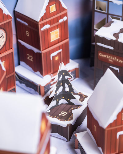 Ein festlich verschneites Pappdorf mit rotbraunen Häusern und einem großen, mit Schnüren beleuchteten Baum. Die nummerierten Fenster offenbaren Überraschungen im Pflanzen Adventskalender (Dschungel-Version) - perfekt für Zimmerpflanzen-Liebhaber.