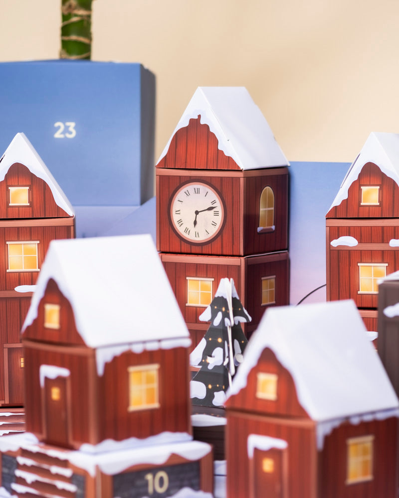 Eine Nahaufnahme eines festlich geschmückten Dorfes zeigt Modellhäuser mit verschneiten Dächern, leuchtenden Fenstern und kleinen Blumentöpfen, die den winterlichen Charme des Pflanzen-Adventskalenders heraufbeschwören.