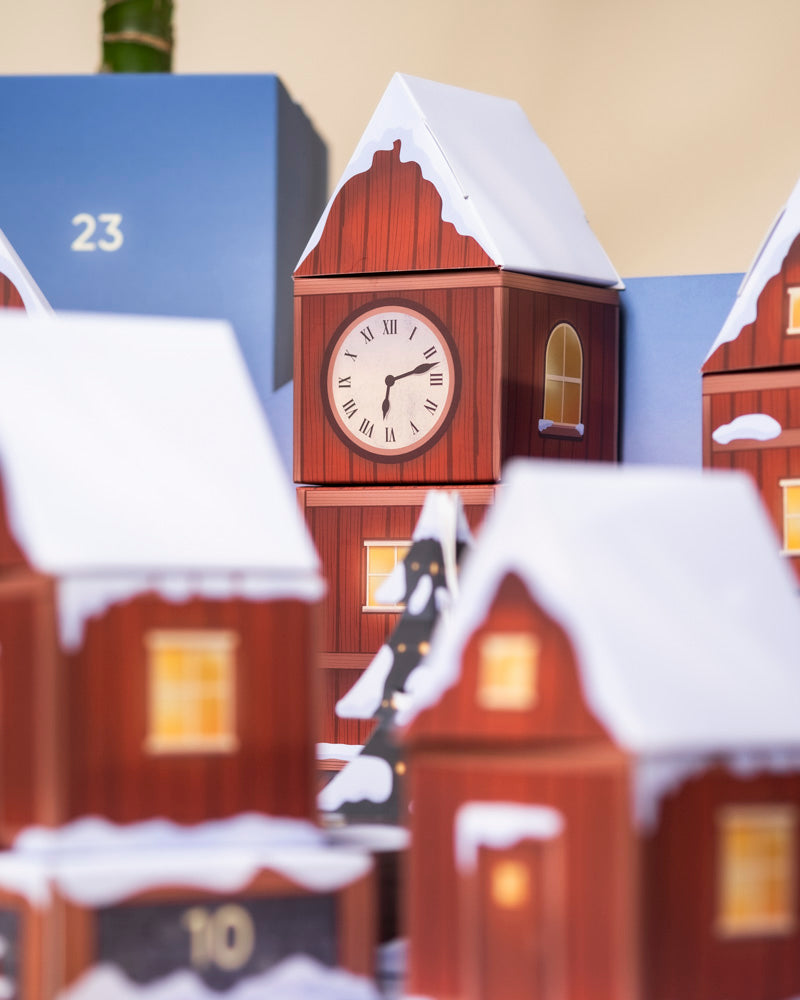 Ein festliches Miniaturdorf mit roten Häusern, verschneiten Dächern und einer Turmuhr um 10:10 Uhr leuchtet warm. Im Hintergrund deutet ein blaues Gebäude mit der Aufschrift "23" auf den Pflanzen-Adventskalender (Dschungel-Version) hin.