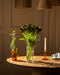 Auf einem runden Holztisch stehen eine gläserne Lieblingsvase mit Alstroemeria rot (10 Stk.), eine kleine bernsteinfarbene Vase mit Grünzeug, ein bernsteinfarbener Kerzenleuchter mit einer weißen Kerze und weiße Teetassen. Warmes Licht und Korblampen schaffen ein gemütliches Ambiente.