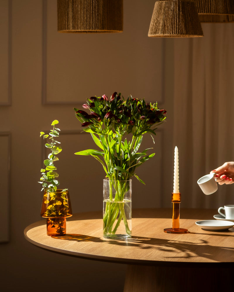 Auf einem runden Holztisch stehen eine mit Alstroemeria rot (10 Stk.) gefüllte Lieblingsvase, eine kleine bernsteinfarbene Vase mit Grünzeug, ein bernsteinfarbener Kerzenständer mit einer weißen Kerze und eine Hand, die aus einem kleinen weißen Krug gießt. Die warme Beleuchtung schafft eine gemütliche Atmosphäre.