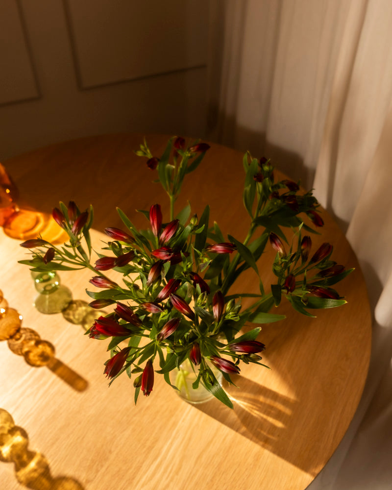 Zehn rote Alstroemeria (Alstroemeria rot, 10 Stk.) mit grünen Blättern in Ihrer Lieblingsvase stehen auf einem runden Holztisch im Sonnenlicht, akzentuiert durch dekorative Glasperlen und eine kleine Bernsteinflasche für ein gemütliches Ambiente.