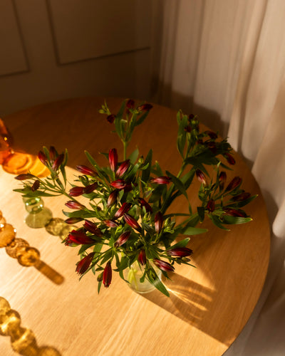 Zehn rote Alstroemeria (Alstroemeria rot, 10 Stk.) mit grünen Blättern in Ihrer Lieblingsvase stehen auf einem runden Holztisch im Sonnenlicht, akzentuiert durch dekorative Glasperlen und eine kleine Bernsteinflasche für ein gemütliches Ambiente.