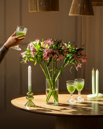 Eine Hand hält ein Glas mit einem grünen Getränk über einem runden Holztisch mit einer Vase mit frischen Blumen, darunter Alstroemeria rosa (10 Stk.), zwei weiteren Gläsern und dekorativen Kerzenhaltern. Warmes Licht schafft eine gemütliche Atmosphäre.