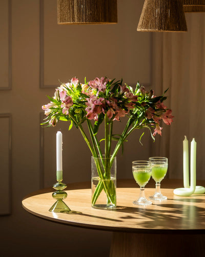 Eine klare Glasvase mit Alstroemeria rosa (10 Stk.) steht auf einem runden Holztisch, daneben eine brennende weiße Kerze in einem grünen Halter, zwei grüne Getränke in eleganten Gläsern und zwei nicht brennende grüne Kerzen. Warmes Licht taucht die Szene.