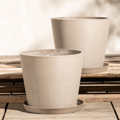 Zwei beigefarbene, minimalistische Pflanzkübel-Set Amsterdam (2x 30) mit passenden Untersetzern stehen auf einer Holzoberfläche. Die Töpfe sind zylindrisch mit leicht konischer Form und die Untersetzer sitzen jeweils direkt darunter. Sie bestehen aus Recycling-Kunststoff und verfügen über ein effizientes Drainagesystem. Den Hintergrund bildet eine schlichte, helle Wand.