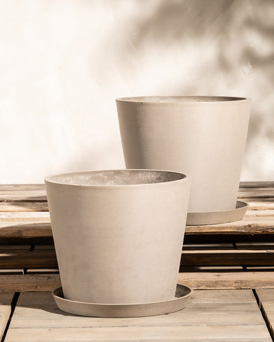 Zwei beige, minimalistisch anmutende Pflanzkübel-Set Amsterdam (2x 30) stehen auf einer Holzoberfläche. Die Töpfe sind rund, verjüngen sich zum Boden hin leicht und ruhen auf passenden Untersetzern. Sie sind aus langlebigem Recycling-Kunststoff gefertigt und verfügen über ein integriertes Drainage-System. Der Hintergrund ist eine helle, neutrale Wand mit weichen Schatten und durchscheinendem Sonnenlicht.