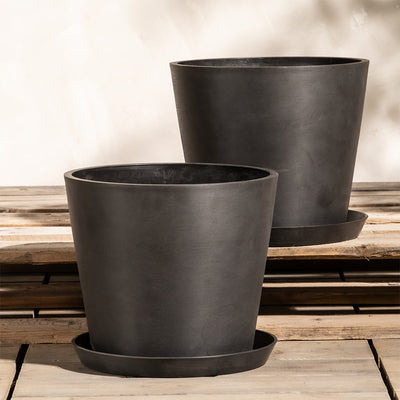 Zwei große, mattschwarze, zylindrische Pflanzkübel-Set Amsterdam (2x 30) mit leicht konisch zulaufenden Unterteilen stehen auf Holzpaletten. Jeder Topf aus Recycling-Kunststoff hat einen passenden Untersetzer darunter. Den Hintergrund bildet eine glatte, helle Wand mit sanften Schattenmustern.
