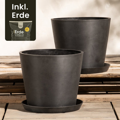Zwei große, schwarze Pflanzkübel-Set Amsterdam (2x 30) mit passenden Untersetzern stehen auf einer Holzfläche. Dahinter ist ein Produktpaket mit der Aufschrift „Inkl. Erde“ teilweise zu erkennen. Das Set-up mit Pflanzkübeln aus Recycling-Kunststoff steht vor einem hellen, unscharfen Hintergrund.