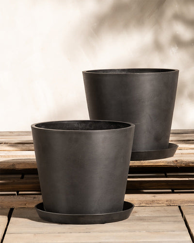 Zwei große, schwarze, zylindrische Pflanzkübel-Set Amsterdam (2x 30) mit passenden Untersetzern stehen auf einer von Sonnenlicht beschienenen Holzfläche. Der Hintergrund zeigt eine helle Wand mit weichen Schatten, die eine warme und einladende Atmosphäre schaffen. Diese Töpfe verfügen über ein innovatives Drainage-System und sind aus Recycling-Kunststoff gefertigt.