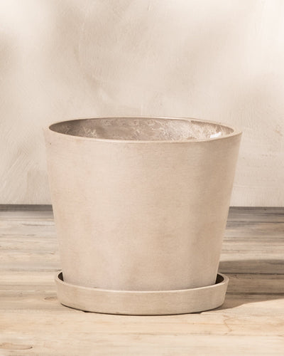 Ein Kunststoff-Pflanzkübel (Amsterdam | 20 cm ⌀) mit passendem Untersetzer steht auf einer Holzoberfläche. Der Topf aus Recycling-Kunststoff hat eine glatte, hellbeige Oberfläche und ein konisches Design. Ein eingebautes Drainagesystem verleiht seinem Stil vor dem strukturierten, cremefarbenen Wandhintergrund Funktionalität.