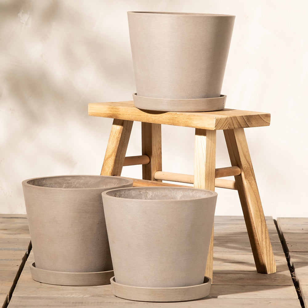 Drei beige Pflanzkübel-Set Amsterdam (3x 20) mit passenden Untersetzern sind kunstvoll arrangiert. Zwei stehen auf einem Holzhocker, während der dritte davor auf dem Boden steht. Der Hocker ist in Naturholzoptik und mit Lattendesign gestaltet. Der Hintergrund ist eine cremefarbene Wand, die für einen minimalistischen Touch sorgt.