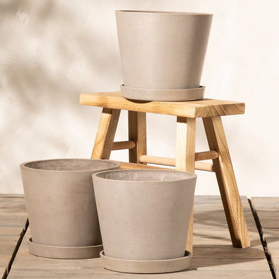 Drei beige Pflanzkübel-Set Amsterdam (3x 20) mit passenden Untersetzern sind kunstvoll arrangiert. Zwei stehen auf einem Holzhocker, während der dritte davor auf dem Boden steht. Der Hocker ist in Naturholzoptik und mit Lattendesign gestaltet. Der Hintergrund ist eine cremefarbene Wand, die für einen minimalistischen Touch sorgt.