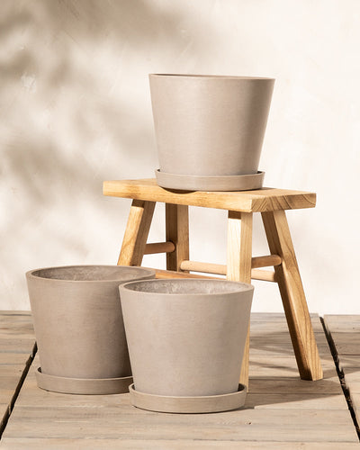 Drei beige Pflanzkübel-Set Amsterdam (3x 20) sind in einer stufenförmigen Anordnung angeordnet: einer auf einem kleinen Holzhocker und zwei auf dem Boden. Der Hintergrund ist neutral, und Licht wirft Schatten, wodurch eine warme, einladende Atmosphäre entsteht. Diese Pflanzkübel sind aus Recycling-Kunststoff gefertigt und verfügen über ein effizientes Drainagesystem für optimale Pflanzengesundheit.