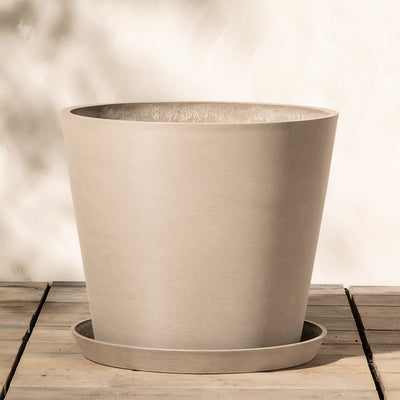 Ein großer, leerer, beiger Keramiktopf steht auf einer Holzoberfläche. Der Kunststoff-Pflanzkübel (Amsterdam | 40 cm ⌀) hat einen passenden Untersetzer darunter und steht vor einem neutralen, leicht strukturierten Hintergrund. Schatten von nahe gelegenen Objekten erzeugen einen weichen, gesprenkelten Lichteffekt auf der Szene.