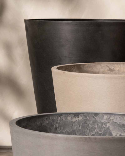 Nahaufnahme von drei zylindrischen Pflanzkübeln vom Typ Kunststoff Pflanzkübel (Amsterdam | 20 cm ⌀) mit glatter Oberfläche. Sie sind versetzt angeordnet und weisen unterschiedliche Farbtöne von Dunkelgrau bis Beige auf. Schatten und Licht spielen auf den strukturierten Oberflächen und erzeugen eine minimalistische und moderne Ästhetik, während sie gleichzeitig über ein innovatives Entwässerungssystem verfügen.