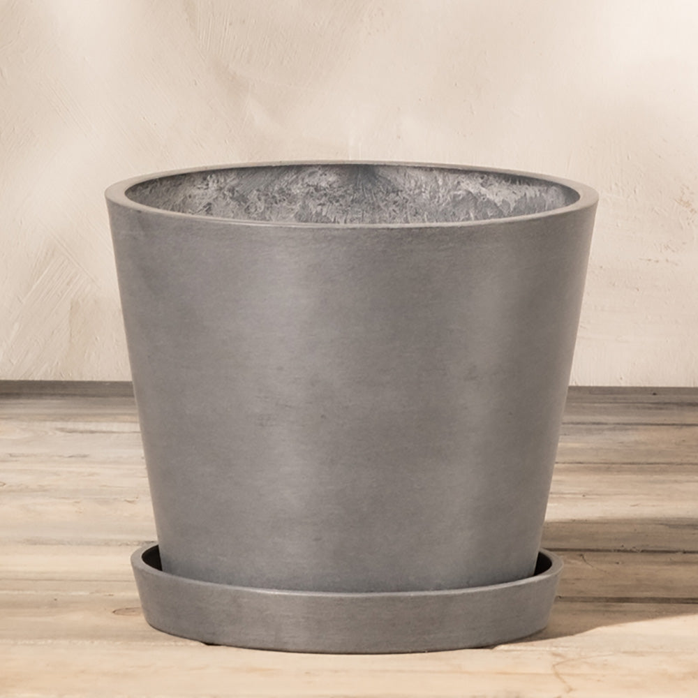 Ein grauer, zylindrischer Pflanzkübel aus Kunststoff (Amsterdam | 20 cm ⌀) steht auf einem passenden Untersetzer. Der Topf hat ein schlichtes, minimalistisches Design mit glatter Textur und ist aus Recycling-Kunststoff gefertigt. Er verfügt über ein effizientes Drainagesystem und steht vor einem neutralen Hintergrund auf einer Holzoberfläche.