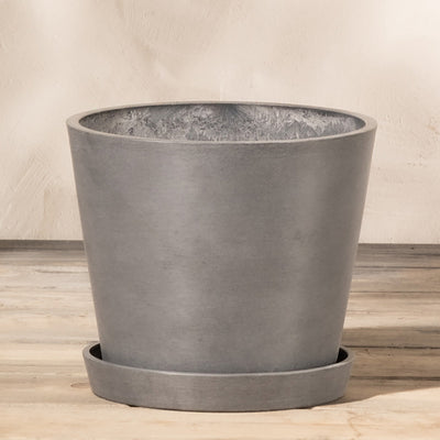 Ein grauer, zylindrischer Pflanzkübel aus Kunststoff (Amsterdam | 20 cm ⌀) steht auf einem passenden Untersetzer. Der Topf hat ein schlichtes, minimalistisches Design mit glatter Textur und ist aus Recycling-Kunststoff gefertigt. Er verfügt über ein effizientes Drainagesystem und steht vor einem neutralen Hintergrund auf einer Holzoberfläche.