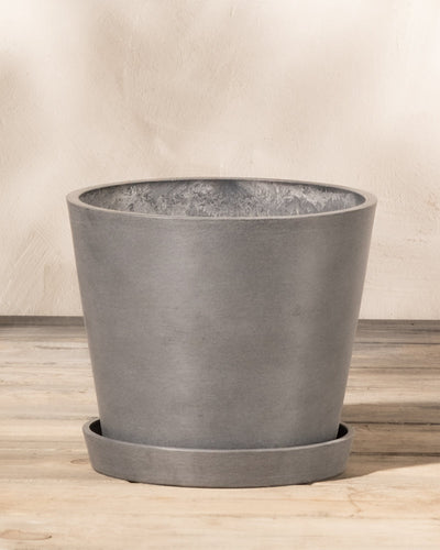 Ein schlichter, moderner, grauer Kunststoff-Pflanzkübel (Amsterdam | 20 cm ⌀) von EcoPots steht auf einem passenden Untersetzer vor einem hellbeigen Hintergrund. Der aus Recycling-Kunststoff gefertigte Topf ist leer und steht auf einer Holzoberfläche. Das Gesamtdesign ist schlicht und minimalistisch.