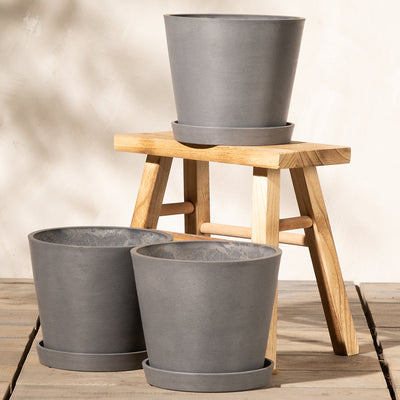 Ein kleiner Holzhocker mit vier Beinen trägt ein graues Pflanzkübel-Set Amsterdam (3x 20) mit passendem Untersetzer. Vor dem Hocker stehen zwei weitere graue Pflanztöpfe mit passenden Untersetzern auf einer Holzfläche. Diese Töpfe aus Recycling-Kunststoff verfügen über ein effizientes Drainage-System. Den Hintergrund bildet eine helle Wand, wodurch eine schlichte und elegante Szene entsteht.